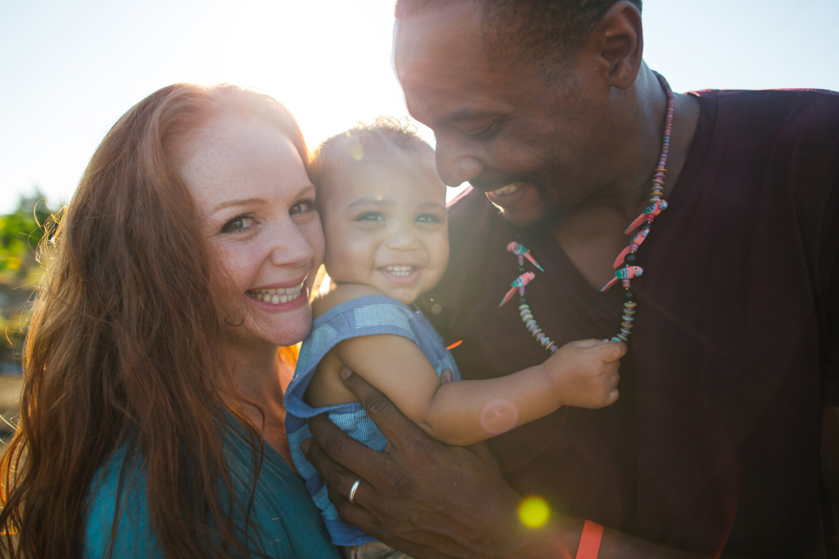 Young Happy Family Of Three Enjoying Each Other Outside On Sunny Day