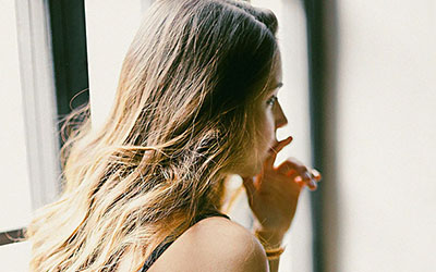 A woman sits near a bright window, looking thoughtful with her hand near her mouth.