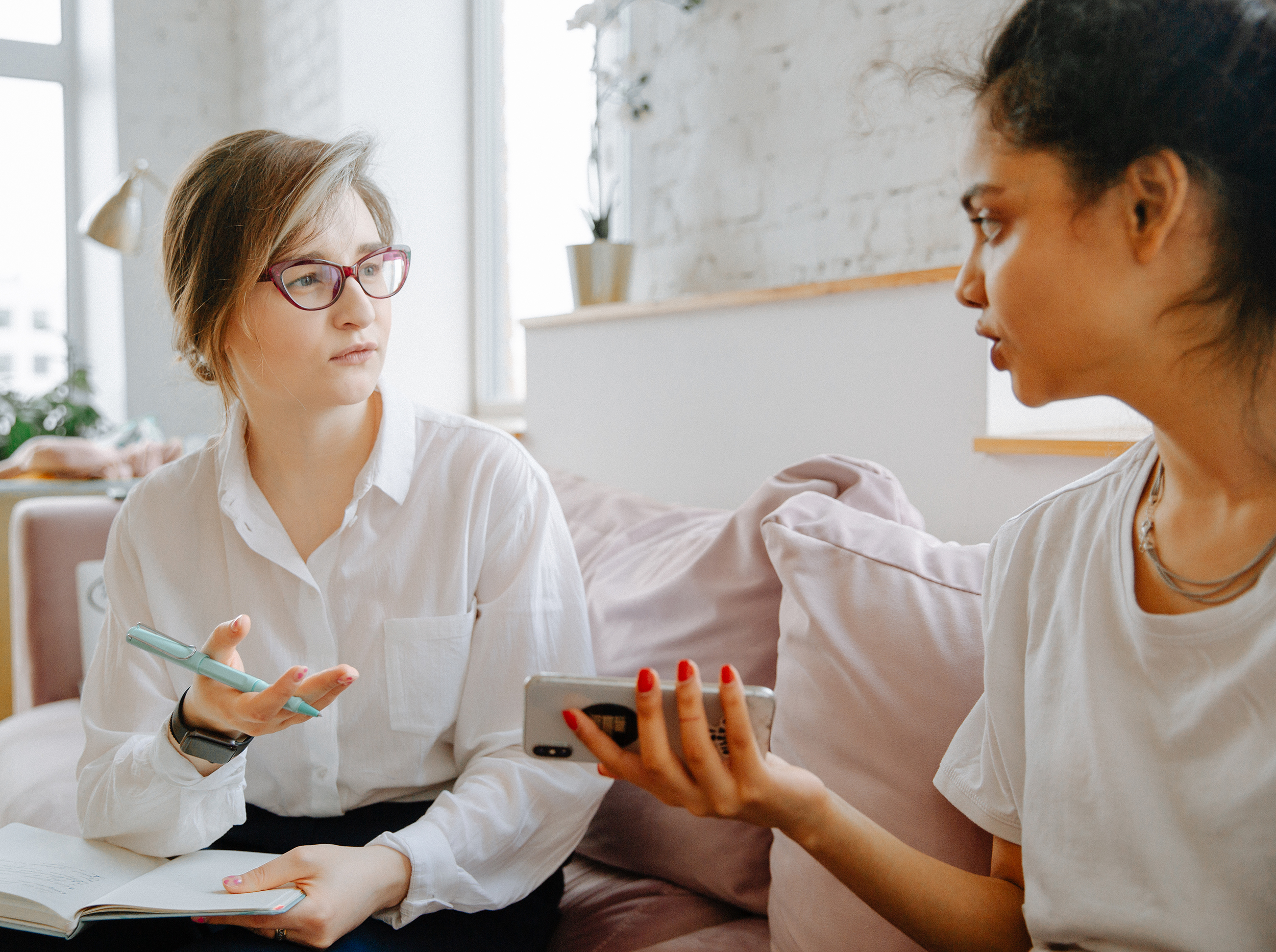 Two women sit on a couch in a bright room, engaged in conversation. One holds a notebook and pen while listening, and the other gestures with a smartphone in her hand.