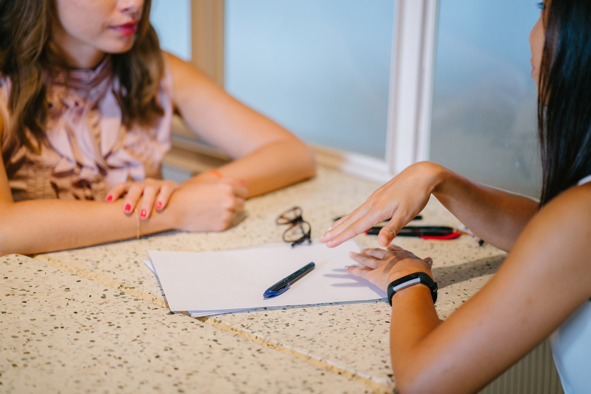 Two people sit at a table in conversation, with papers, a pen, and glasses between them. One person gestures with their hand while the other listens.
