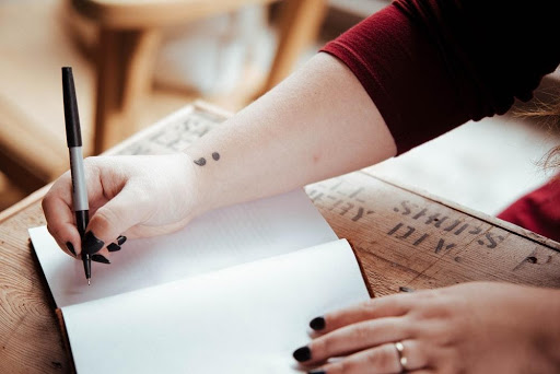 Close up of a person writing in a notebook at a wooden table, with a small semicolon tattoo visible on their wrist.